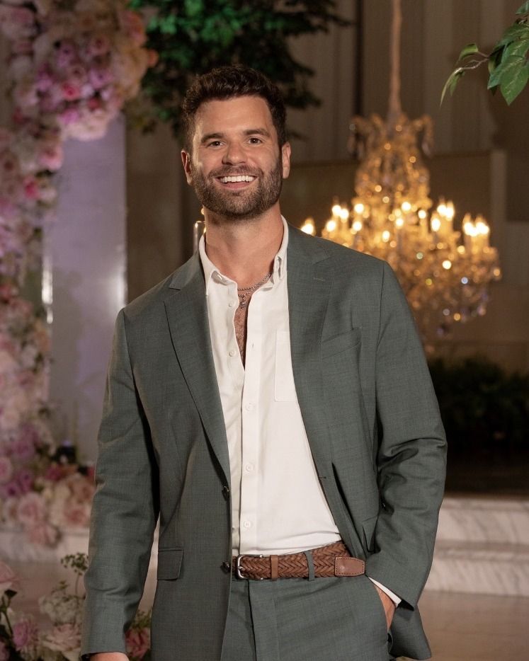 Nick Uhlenhuth in a green suit jacket, smiling, in front of a chandelier and a wall of pink flowers